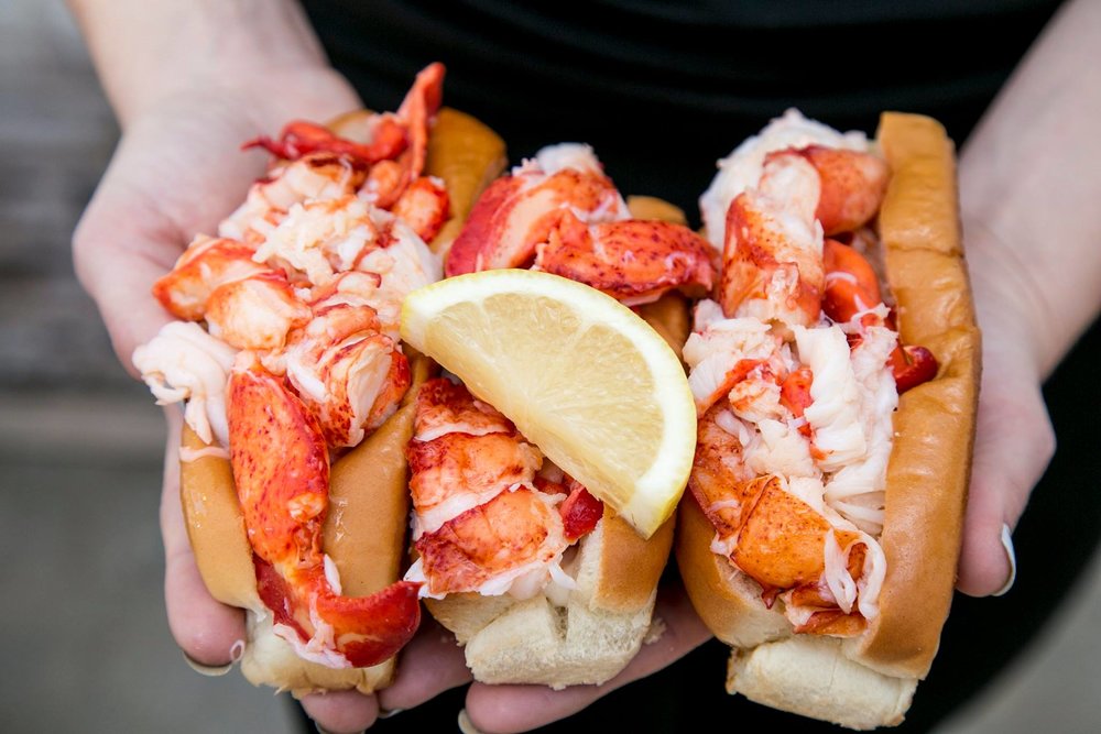 Vendor holding up three fresh lobster rolls, the kind of event photo that promotes a truck